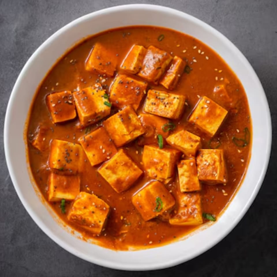 Indian paneer curry with cubes of cheese in a rich orange sauce, garnished with seeds and herbs.